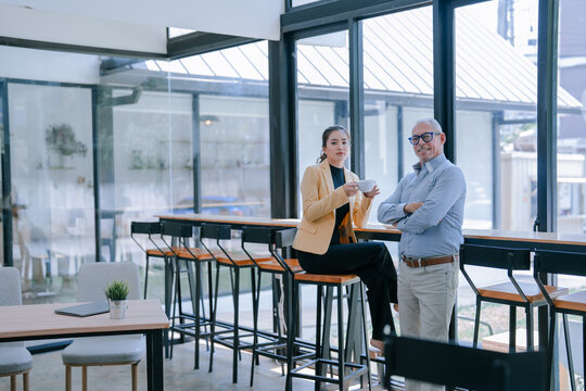 Two business partners, a woman sitting on a stool and holding a cup of coffee and a man standing with his arms crossed, are taking a break in a modern office cafeteria with large windows