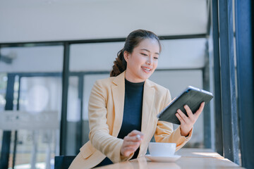 Young asian businesswoman is using tablet and drinking coffee during break in cafe, she is smiling and looking at tablet while stirring coffee