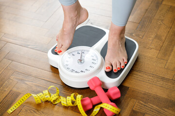 Young woman stepping on scale with dumbbells and measuring tape on wooden floor