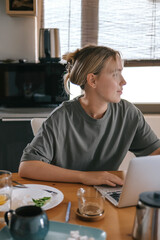  Gen Z woman working from home at cozy kitchen table with laptop and phone. Digital nomad...