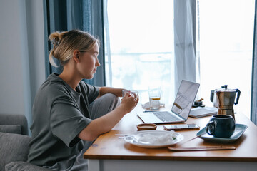  Gen Z woman working from home at cozy kitchen table with laptop and phone. Digital nomad...