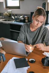  Gen Z woman working from home at cozy kitchen table with laptop and phone. Digital nomad lifestyle, mindfulness, remote work, balance, mental calm and modern freelancing