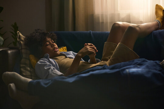 Young person lying on couch with smartphone, wearing casual clothes and absorbing in an activity on the device, creating a comfortable indoor setting without daylight - Powered by Adobe