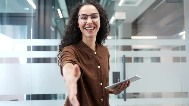 Happy young businesswoman extending her hand to say hello while standing in modern business office. Smiling positive female manager in glasses with digital tablet in hands looking friendly at camera