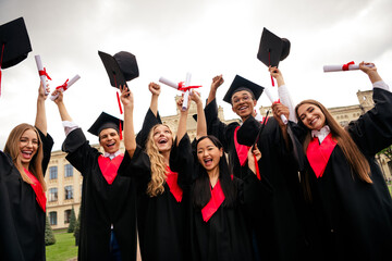 Joyful group of young graduates celebrating outdoors, wearing caps and gowns, holding diplomas...