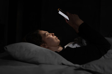 Low-light photo showing a woman reading from a phone while lying on her back in bed. The screen...