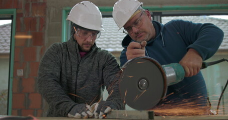 Two construction workers cutting metal with electric grinder, intense sparks flying, safety gear...