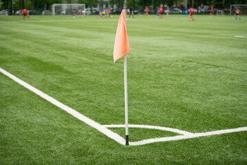 A vibrant corner flag stands at the edge of a manicured green soccer field. Players from two teams engage in a competitive match in the background, showcasing skillful play and teamwork © Oleksandr Zinchenko