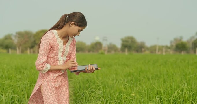 Indian young adult farmer woman lady wear casual dress desi life outdoor holding tablet device website looking screen asian smiling teen age girl use tab yield data outside work rural area day time