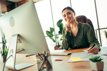 Smiling young professional seated at desk in modern office, working with confidence among green...