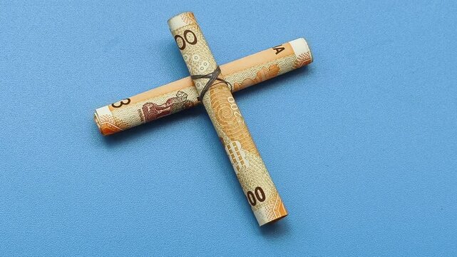 Hand placing a cross symbol made with currency notes on blue table surface with copy space. Closeup top view. Tithe, church donation, charity, contribution, religious tax and financial aid concept.
