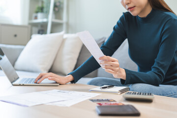 Stressed asian young business woman, employee using laptop computer to calculate expenses, hand holding bills and receipt for to payment on table at home. Financial, finance of banking concept.