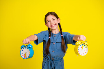 Smiling girl holding colorful alarm clocks in both hands against a vibrant yellow background