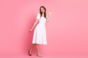 Cheerful young woman in elegant white dress waving hello against vibrant pink background,...