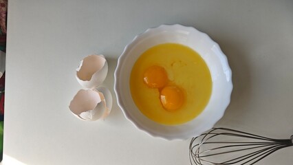Two raw eggs in a white bowl with broken eggshells and a metal whisk on a light background. Concept...