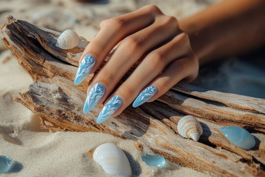 A woman's hand with long blue and white decorative nails rests on a piece of driftwood surrounded by seashells on sandy beach