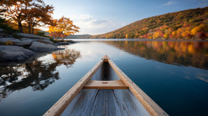 Naklejka premium A wooden canoe floats on a calm lake surrounded by autumn trees and distant hills under a bright sky.