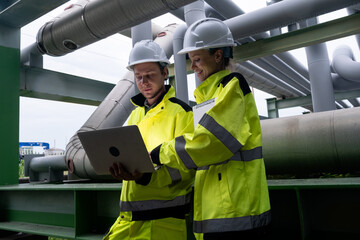 Engineers collaborate on a project at an industrial site during daylight hours wearing protective gear and using a laptop