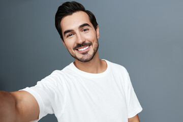 Friendly smiling young man with dark hair wearing a casual white t-shirt, posing for a selfie against a solid grey background. Lifestyle and modern concept.