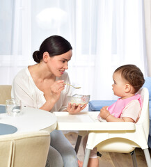 Mother feeding baby with a spoon. Spoon-feed. Happy young mother with a baby in the kitchen interior. Fresh vegetables and fruits.