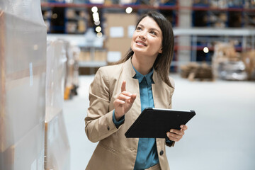 A confident businesswoman using a tablet while assessing inventory in a modern warehouse environment showcasing professionalism.