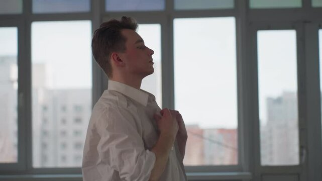 side view of young man adjusting white shirt with focused expression in bright room, standing near large window with blurred urban background, casual movement during quiet indoor moment