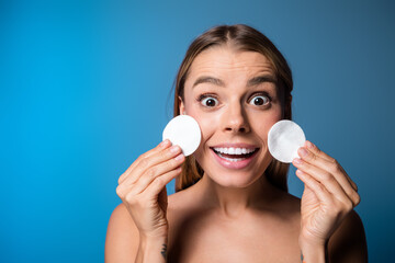 Young woman with cotton pads on blue background expressing happiness and concept of skincare and...