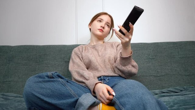 Beautiful teen girl sitting on sofa in living room, using remote control, changing channels, enjoying tv programs in a cozy home environment
