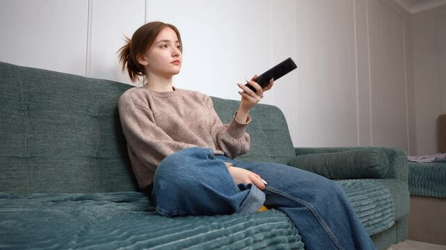 Beautiful teen girl sitting on sofa in living room, using remote control, changing channels, enjoying tv programs in a cozy home environment