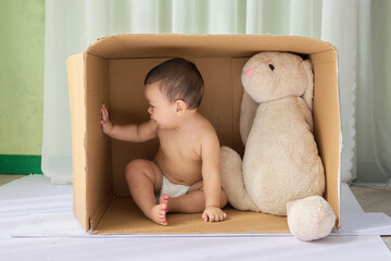a baby sitting in a cardboard box with a teddy bear beside them. The background of the image is a...