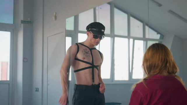 rear view of woman with golden hair seated facing man removing belt looping it around neck then casting aside during seductive dance in mirrored room spotlighting harness sunglasses baton cloth props