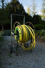 A sturdy garden hose reel with yellow and green hoses sits on a gravel path, surrounded by lush foliage. The metal frame has wheels for easy transport, perfect for efficient watering and irrigation