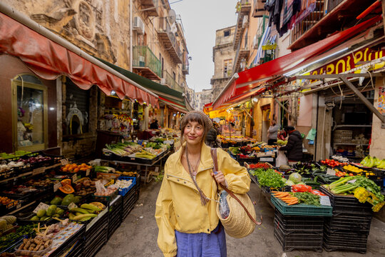 A woman in a yellow jacket strolls through a vibrant local market in Sicily, surrounded by fresh produce. The image conveys authentic travel, cultural immersion, and slow lifestyle 