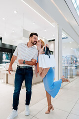 Happy young couple sharing a kiss while joyfully holding shopping bags, strolling through a mall after purchasing elegant jewelry from a luxurious store