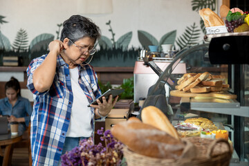 Asian senior woman in plaid shirt holding tablet while scratching neck standing in front of bakery display case with concerned expression showing mistake or confusion while checking inventory in cafe