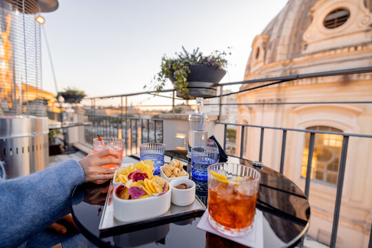Elegant aperitivo with cocktails and snacks on a Roman rooftop at sunset. A hand reaches for a drink as the dome glows in warm evening light