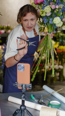 Fototapeta premium Florist conducts online training on making bouquets using a phone placed on a tripod on a table. Florist records an educational video. Small business. Close-Up. She demonstrates the pruner she uses.