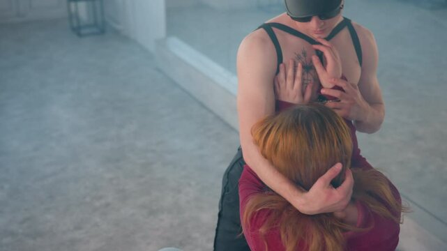 man on ground caressing woman hand to bare chest with symbol tattoo while handcuff hangs from belt beside harness straps in bright mirrored dance studio filled with soft natural light