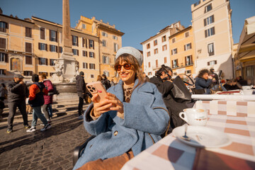 Woman in a blue coat scrolls her phone with a smile at a café table in a vibrant Roman square,...