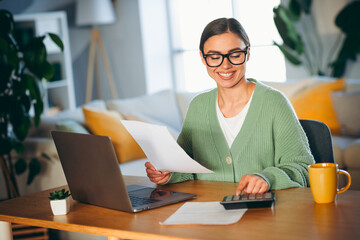 Young woman wearing green sweater working from home with a laptop and calculator in a cozy living...