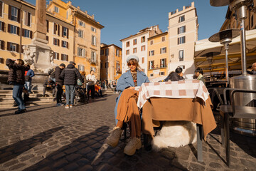 Smiling woman in a blue coat enjoys her coffee in a sunlit Roman square, surrounded by warm-toned...