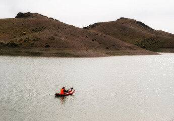 Male tourist in life jacket on red inflatable boat kayak or packraft with paddle floating on water of steppe lake with hills on sunny autumn day