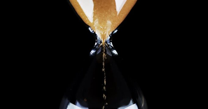 Hourglass. Sands move through hour glass. Sandglass close-up on a black background. Slow motion video. A pile of Golden sand at the bottom of the hourglass, small grains of sand fall from above.