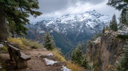 A wooden bench overlooks a breathtaking landscape of snow-covered mountains and lush green trees. Clouds hover above, creating a serene atmosphere in the forest