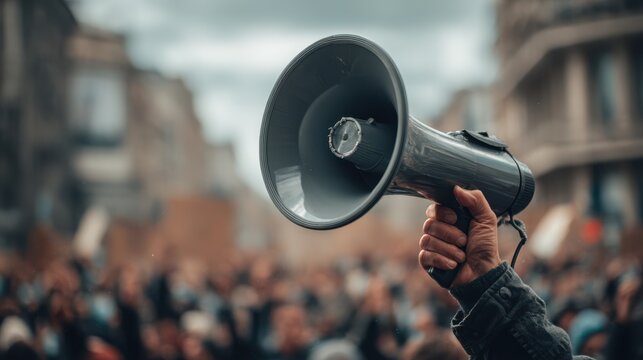 A large crowd is gathered in an urban area while a person holds a megaphone aloft, rallying for a cause during the afternoon. The atmosphere is charged with energy and purpose