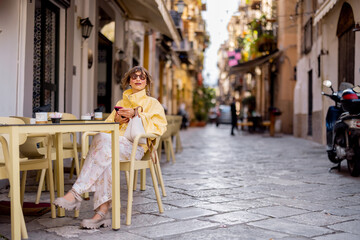 A woman in a yellow jacket checks her phone while sitting at a street cafe in Palermos historic...
