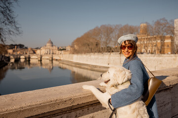 A woman in a blue coat embraces her large white dog on a Roman bridge, gazing toward St. Peters...