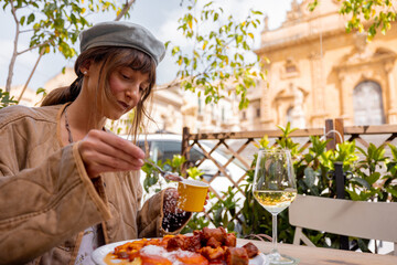 Woman enjoys a plate of traditional Sicilian pasta alla Norma and a glass of white wine at an outdoor cafe in Modica, with Saint Peter Cathedral in the background