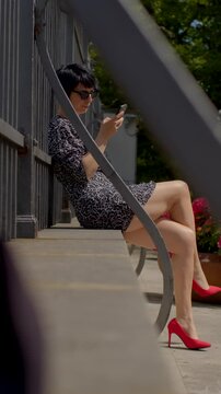 A woman is elegantly seated on the steps, deeply engrossed in her phone while wearing a chic dress and striking red heels