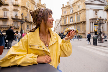 A smiling woman in a yellow jacket enjoys a traditional Sicilian cannolo while sitting at a street...